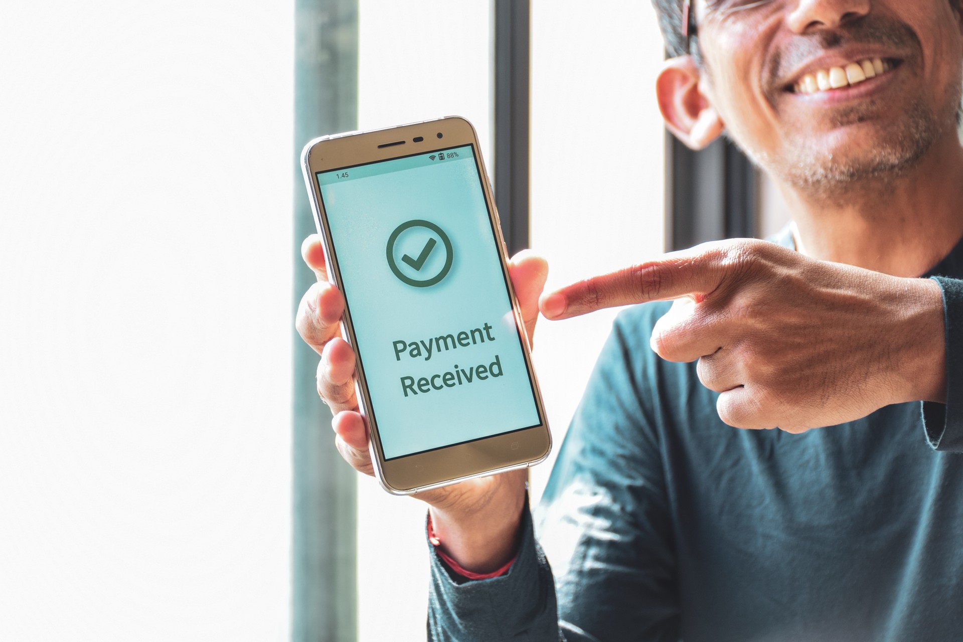 Cropped image of a smiling South Asian man pointing out the notification payment received on his smartphone. Cashless or real time payments concept.